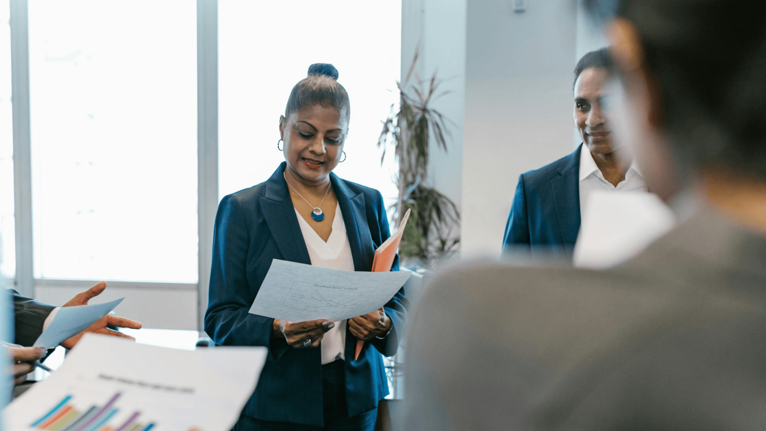 A group of professionals in a business setting, with a focus on a woman leader holding documents and reviewing them. She is dressed in a formal blue blazer with a necklace and is surrounded by colleagues, with some papers and charts in the foreground