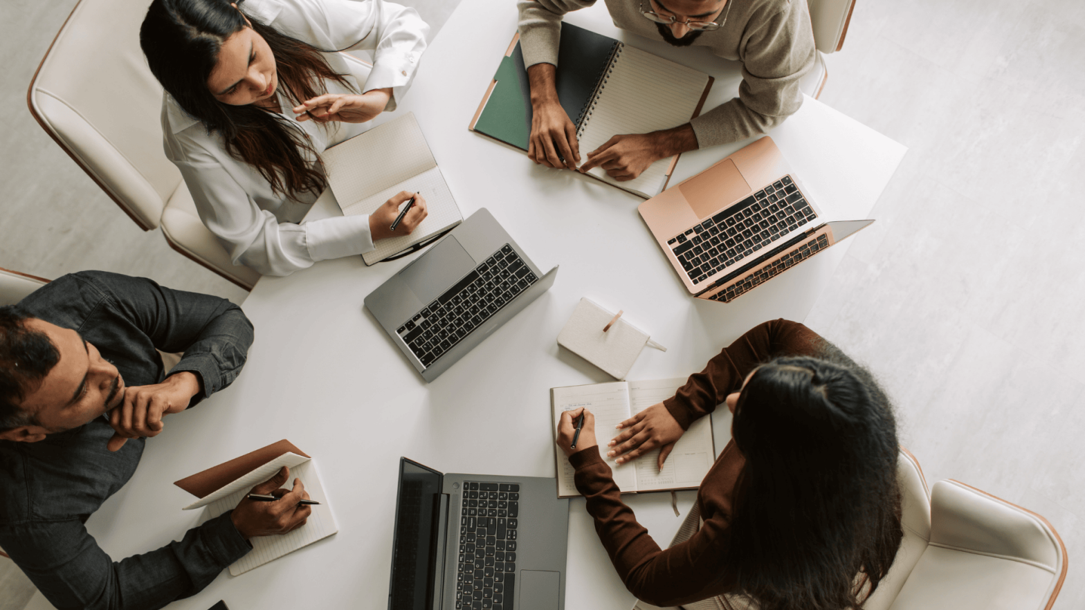 An overhead shot of a collaborative business meeting with four professionals sitting around a table, each with laptops and notebooks. They are engaged in discussion, writing notes, and brainstorming ideas