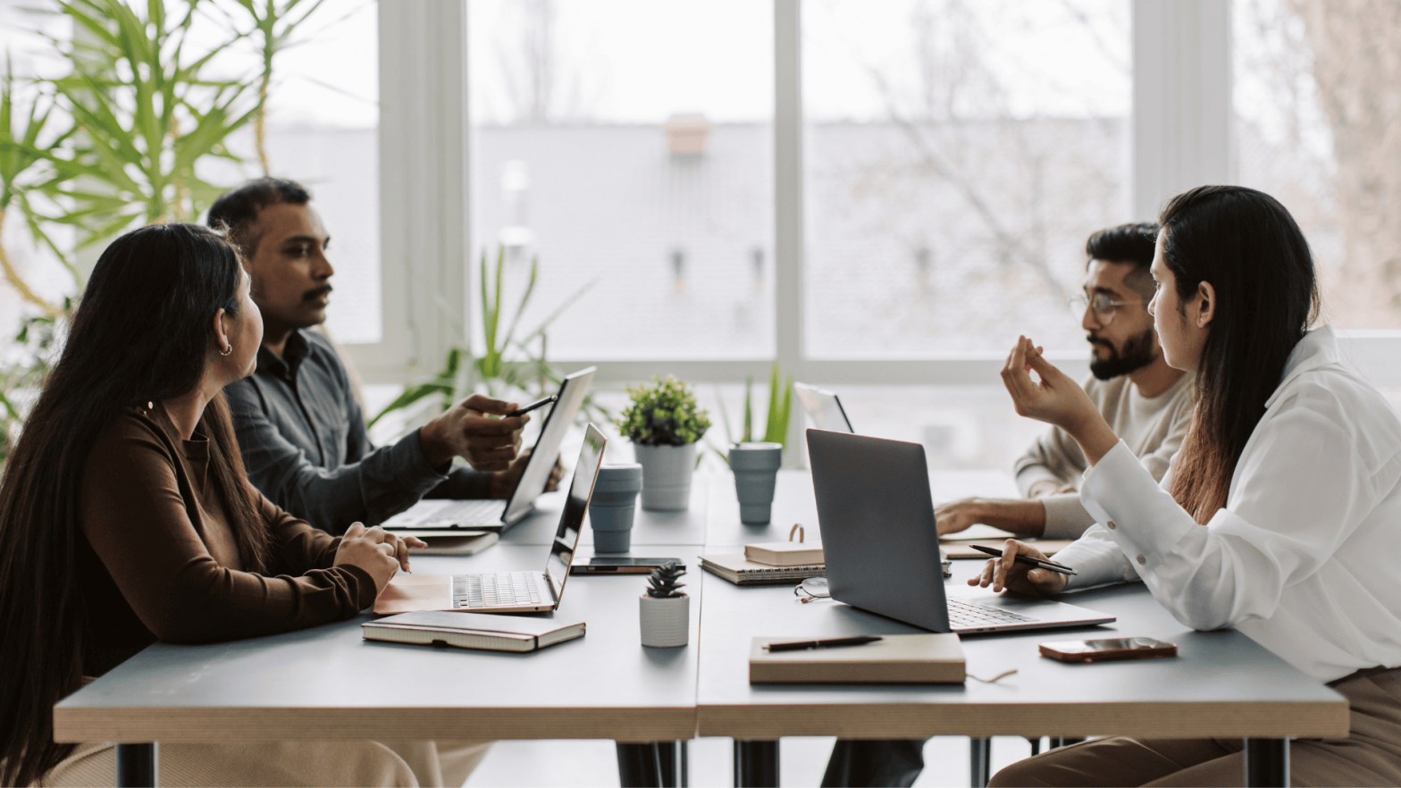 A group of professionals in a modern office space engaged in a meeting. The team, consisting of four people, is seated around a table with laptops, notebooks, and plants. They are discussing and brainstorming ideas, fostering collaboration and innovation in a startup environment
