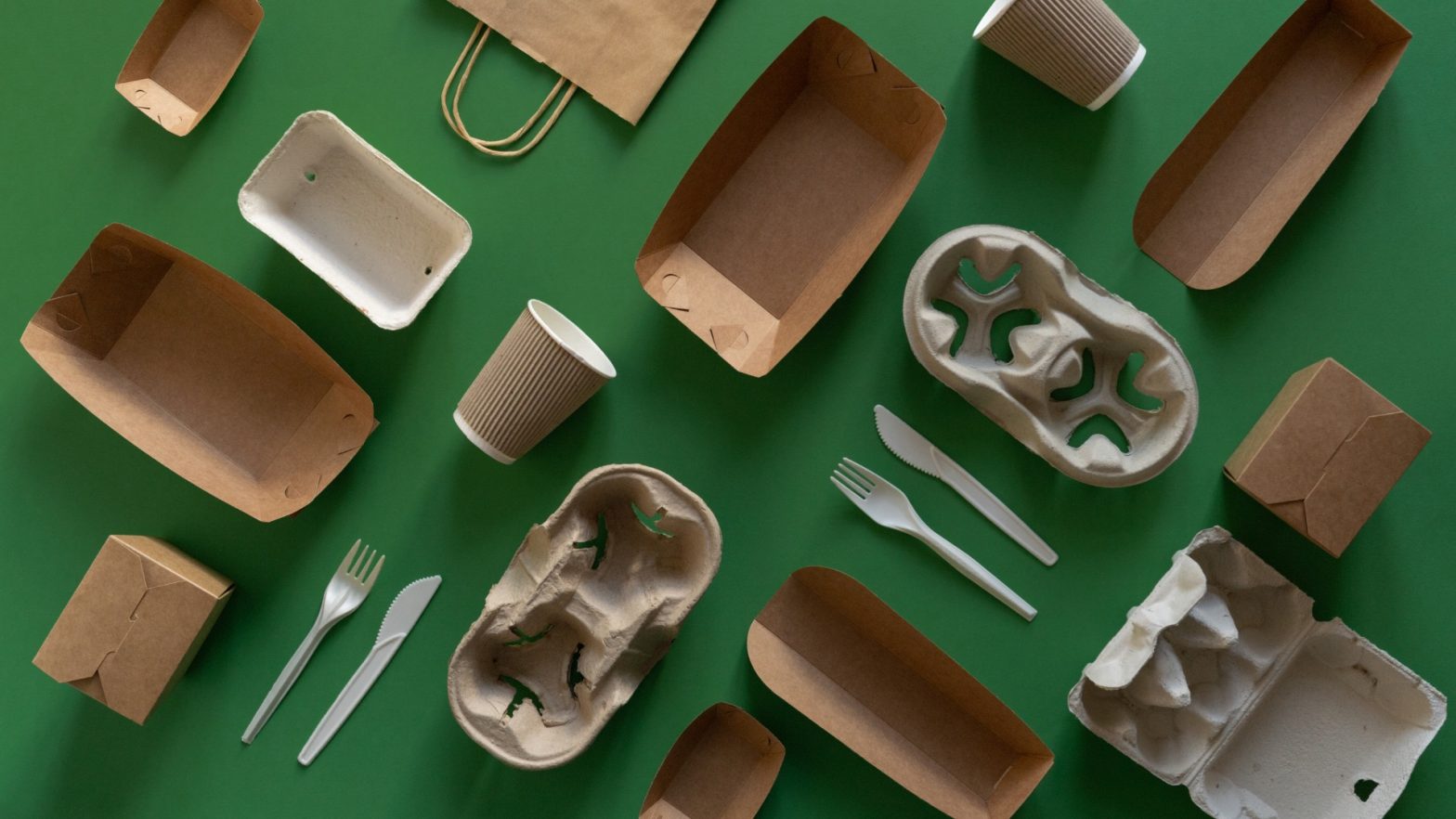 A flat lay of various eco-friendly packaging materials on a green background, including paper boxes, egg cartons, disposable cups, and plastic cutlery