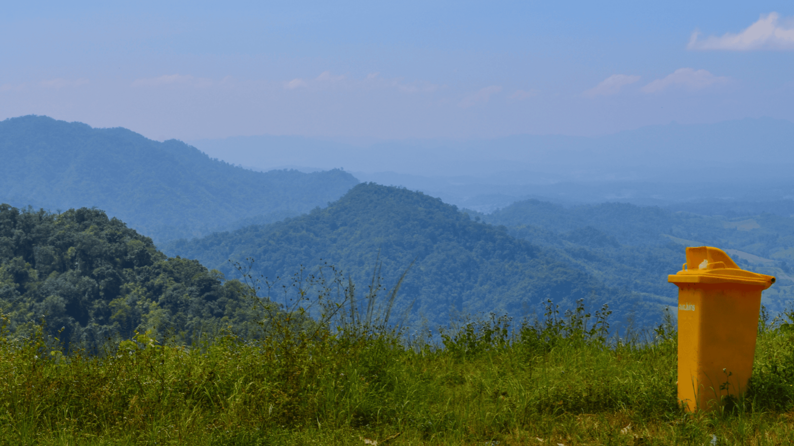 A yellow waste bin sits alone on a grassy hillside, with rolling mountains in the background under a clear blue sky. The image reflects the contrast between waste management efforts and the natural beauty of hill towns
