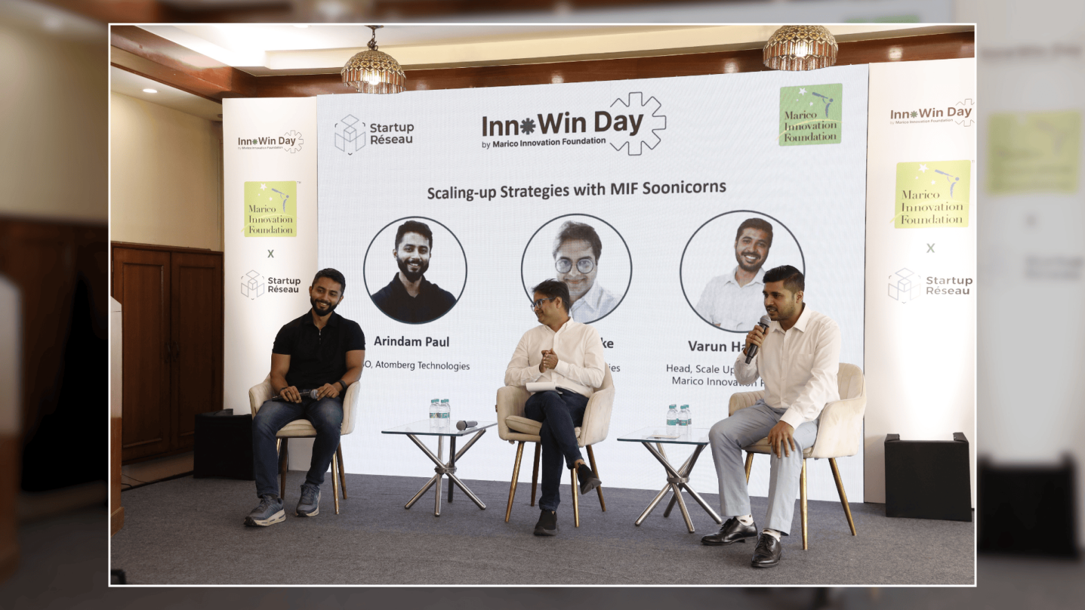 Three men are seated on a stage during a panel discussion at an event called "InnoWin Day" organized by the Marico Innovation Foundation and Startup Réseau. Behind them is a large screen displaying the session title "Scaling-up Strategies with MIF Soonicorns" and their photos and names: Arindam Paul (COO, Atomberg Technologies), Mike (unlisted full name), and Varun Handa (Head, Scale Up Program, Marico Innovation Foundation). The panelists are casually dressed, holding microphones, and engaged in discussion. Two small tables with water bottles are placed between them.