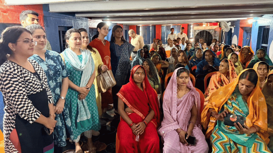 A large group of women, many in colorful saris, gathered in a room, listening attentively to speakers standing on the left.