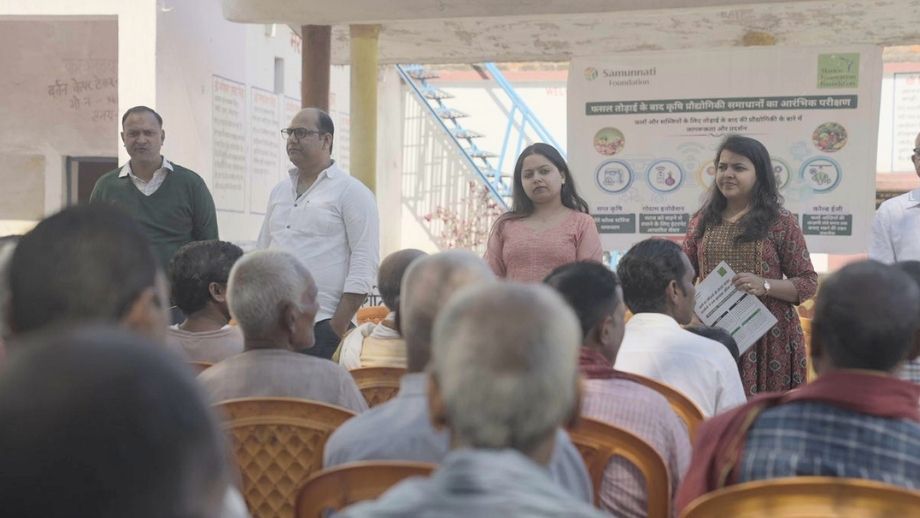 Presenters stand before a seated crowd of farmers in a village setting, explaining new tech initiatives.