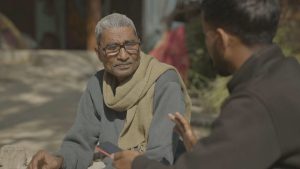 An elderly farmer in a sweater and glasses talks to a person holding a smartphone during a field visit.