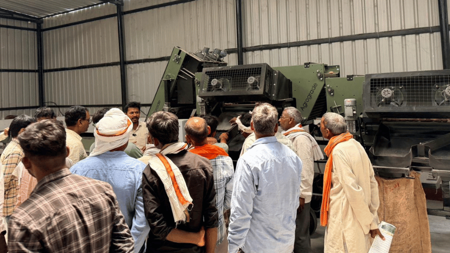A group of farmers closely observing a demonstration of a large, green 'Agrograde' sorting machine inside a facility.