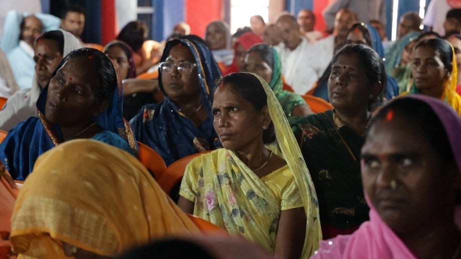 A group of rural women in colorful saris sitting attentively on orange chairs during a community session.