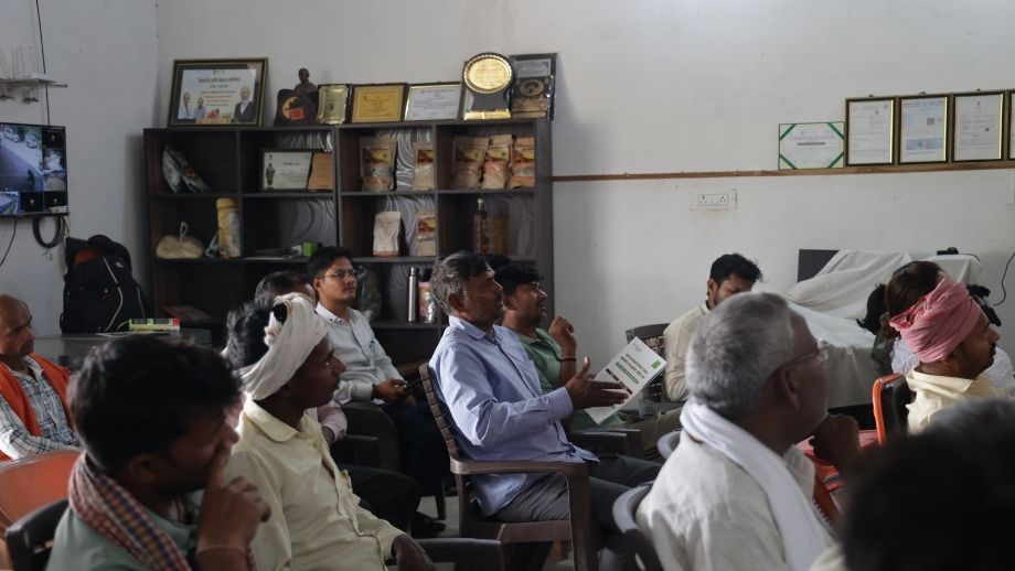 : A group of male farmers seated indoors listening to a session, with one man actively participating while holding a pamphlet.